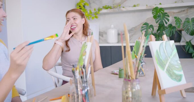 Female friends reacting to brush coming toward face, laughing and painting leaf designs at table