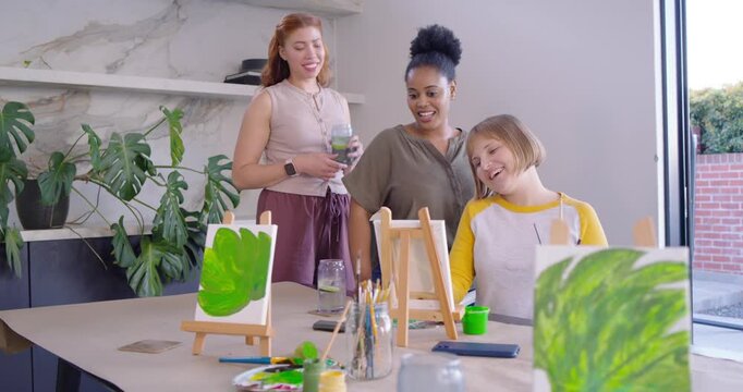 Diverse women friends painting leaf art at table redhead arriving with drink pointing and cheering