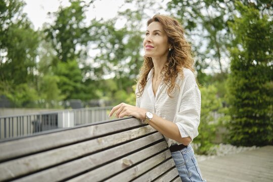 A woman leans against a wooden bench in a lush green park, gazing thoughtfully into the distance. The scene captures the tranquility of a sunny afternoon, surrounded by trees