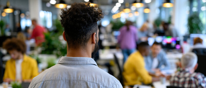 > Rear view of an isolated male employee standing alone watching coworkers collaborate in a modern open-plan office. Concept of workplace exclusion, social isolation, or a newcomer on their first day