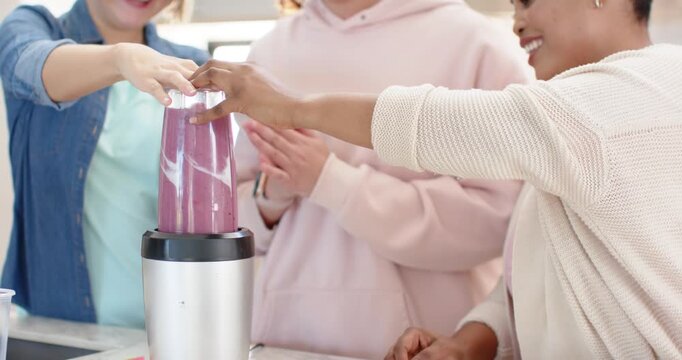 Diverse female friends placing blender cup on base, securing lid, making purple smoothie at counter