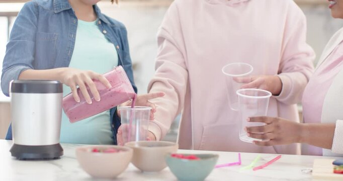 Diverse women in kitchen lifting blender pitcher and pouring pink drink into cups to serve