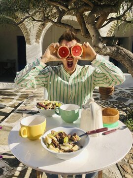 Young man in shirt, grimacing and holding half of a grapfruit as eyes in front of his face, breakfast with muesli and tea on a terrace, Paros, Cyclades Islands, Greece