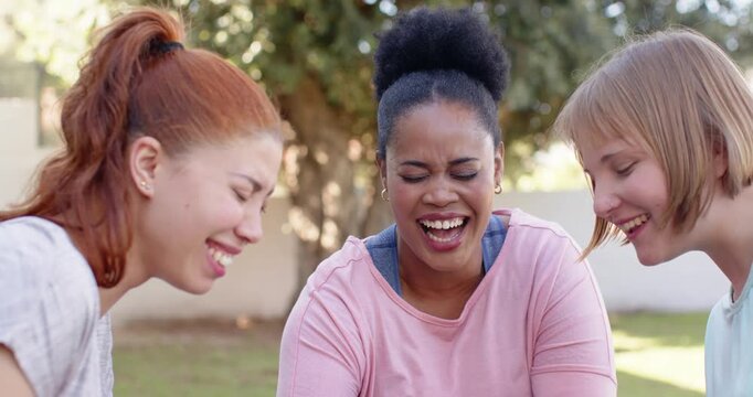 Three female friends spotting unseen object, sitting on grass under tree, pointing and laughing