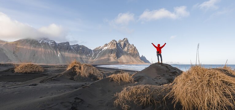 Man in red jacket stretches arms into the air, black lava beach, sandy beach, dunes with dry grass, mountains Klifatindur, Eystrahorn and Kambhorn, headland Stokksnes, mountain range Klifatindur, Austurland, East Iceland, Iceland