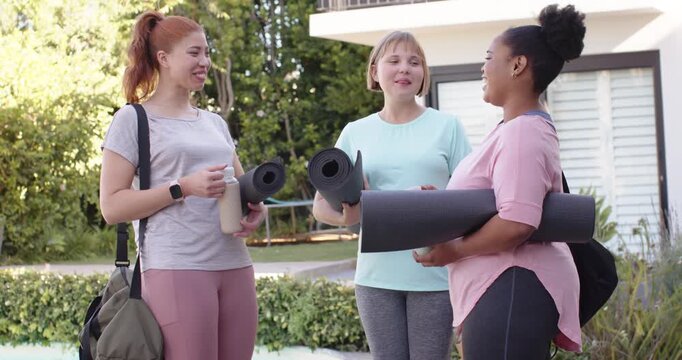 Diverse female friends meeting on wooden deck holding rolled black yoga mats, talking before yoga