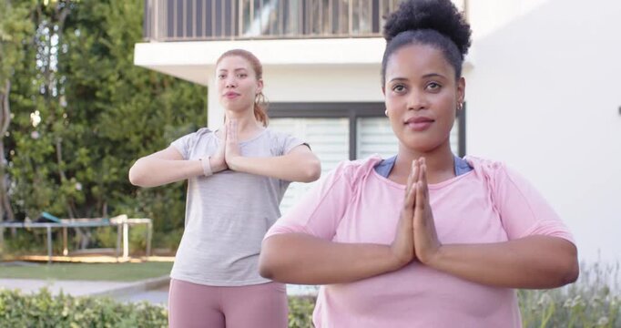 Diverse women in pink grey tops holding prayer hands in yard by trampoline, sunlight causing smiles