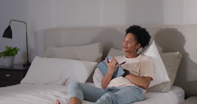 African American man sitting up on bed, lifting blue tablet, using stylus, checking content