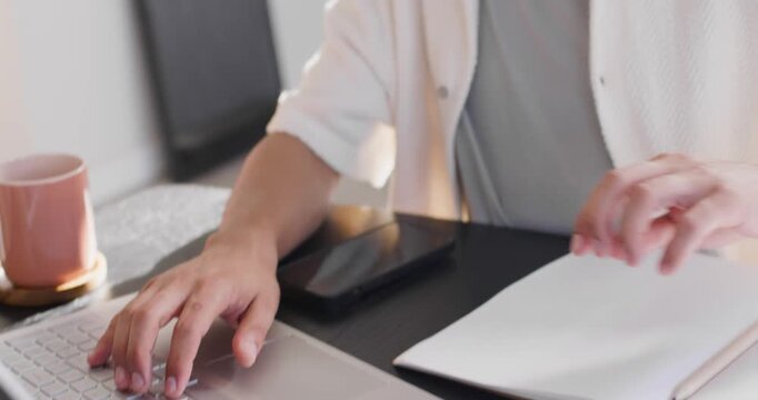 African American man typing on laptop at home, prompted by open notebook and consulting tablet