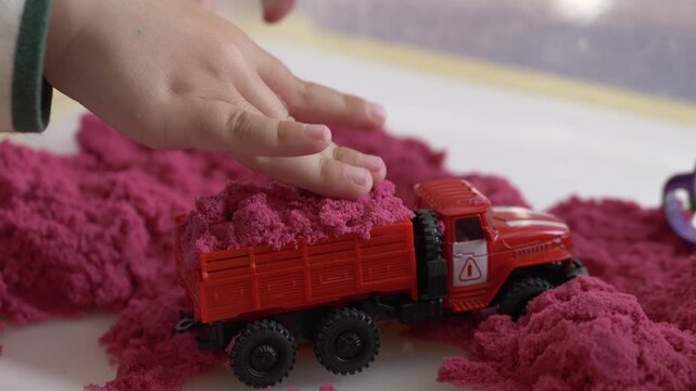 Close-up of toy dump truck loaded with pink kinetic sand. Children's hands explore texture while developing coordination, motor skills and creative problem solving.