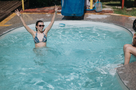 Young woman wearing sunglasses and a patterned bikini raises her arms while standing in a small swimming pool. A carefree summer scene shows friends relaxing and splashing in an outdoor pool.