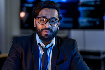 Obraz premium South Asian male network security engineer in a dark blazer and blue badge presents attack vector analysis in a dark room with blue-lit threat dashboards.