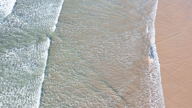 Aerial view of waves rolling onto the sandy shore with white foam and textured water patterns at Perranporth, Cornwall, United Kingdom.