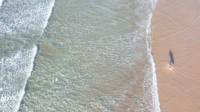 Aerial view of a lone surfer walking along the sandy shore with foamy waves rolling onto the beach in Perranporth, Cornwall, United Kingdom.