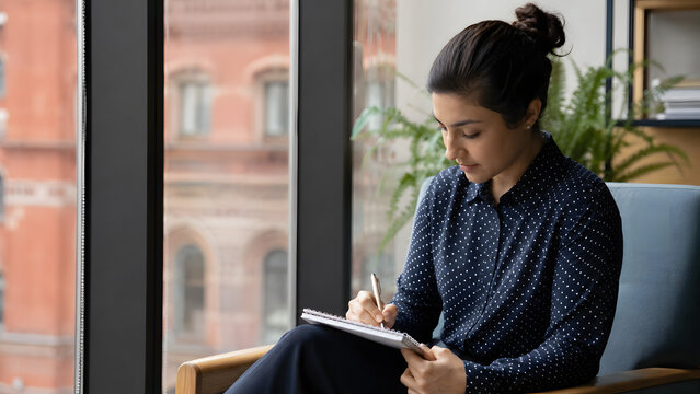 Focused Mindset: A young woman sits in a chair by a large window, deeply engrossed in her work on a tablet. Natural light illuminates her focused expression as she writes.