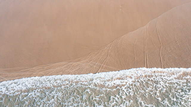 Aerial view of the shoreline where white ocean waves meet the golden sandy beach with water patterns Perranporth, Cornwall, United Kingdom.