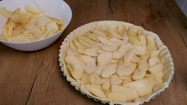 a person sprinkles sugar over apple slices and raisins in a raw pie crust on a wooden table in a home kitchen during baking preparation to sweeten a fruit pie