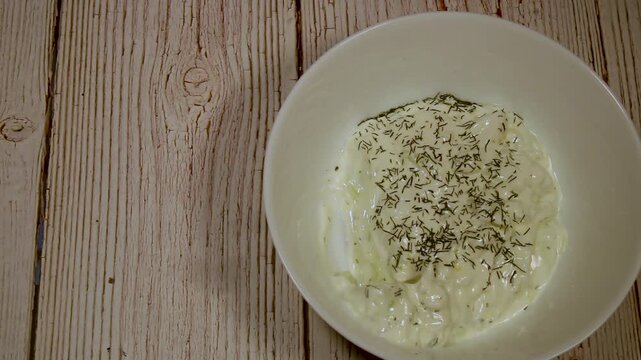 a person dips a fresh cucumber slice into creamy tzatziki herb sauce in a bowl on a wooden table in a home kitchen during food preparation to make a quick snack