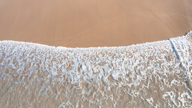 Aerial view of gentle waves breaking on a sandy beach with white sea foam and ripples in the sand in Perranporth, Cornwall, United Kingdom.