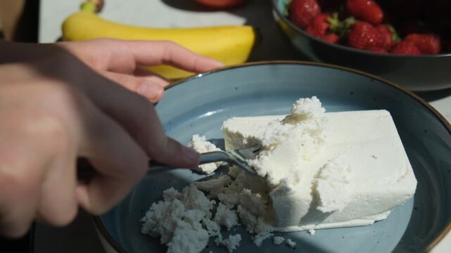 breaking up block of cottage cheese with fork in plate. crumbling cottage cheese with a fork for preparing nutritious breakfast. turning cottage cheese into a crumbly texture before adding sour cream