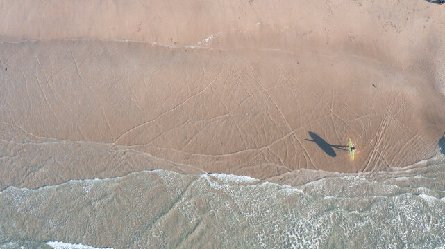 Aerial view of a lone surfer with a yellow board walking along the sandy shore of Perranporth Beach with gentle waves and tracks in the sand Perranporth, Cornwall, United Kingdom.