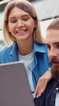 Man and woman sitting on bench in city financial district in casual business style working together on laptop, makes notes, smiling, freelance people, outside