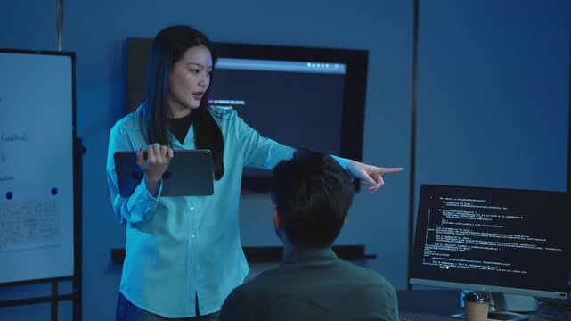 Female project manager holding tablet gesturing while explaining work to male developer, monitors with program code in dark office at night.