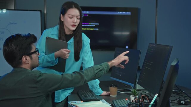 Asian woman with tablet standing while Indian male programmer points at monitor with code, software team solving problem in dark office at night.