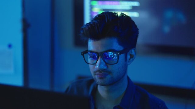 Close up of young Indian man with eyeglasses, computer code reflected in lenses, focused software developer working late in a dark office.