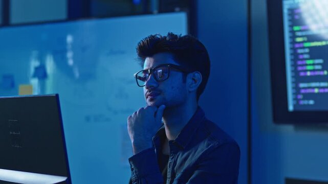 Young Indian man with glasses thinking about solution while looking at monitor, whiteboard and code screen behind, problem solving at night.