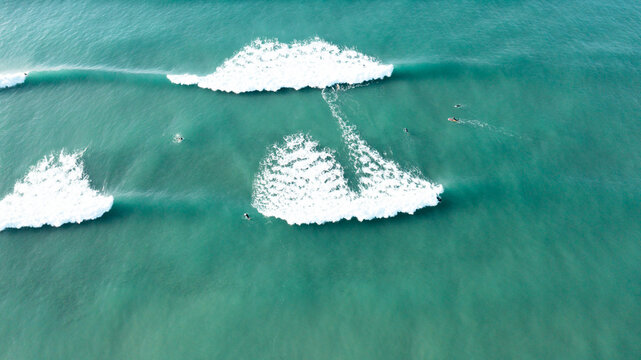 Aerial view of surfers riding turquoise waves with white sea foam and rolling swells in the Atlantic Ocean at Perranporth, Cornwall, United Kingdom.