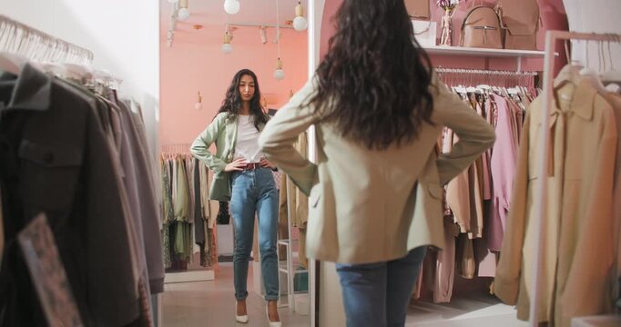 Stylish young woman choosing clothes and comparing outfit options while looking at her reflection in a mirror inside a modern fashion boutique. Female shopper holds hangers with garments and evaluates