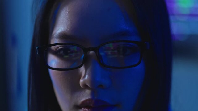 Macro portrait of Asian female programmer wearing eyeglasses with computer code reflection, dark blue light, software development concept.