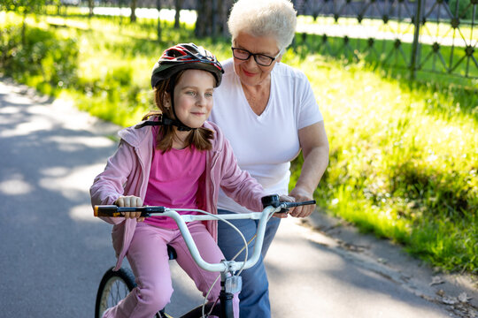 Grandmother helping young granddaughter learn to ride bicycle outdoors. Family support, intergenerational relationship, active aging, confidence building, childhood development. Copyspace