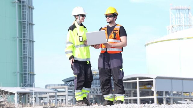 Engineer inspection at industrial plant, safety helmet laptop teamwork as two worker technicians review site audit data near storage tank, outdoor processing maintenance planning at refinery