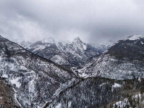Aerial view of snow-capped mountain peaks and a deep valley covered in dense evergreen forests under an overcast sky in CO, United States.