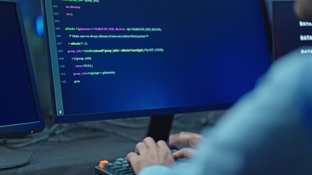 Over shoulder view of programmer's hands on keyboard with colorful source code on screen, software engineering and development at night.