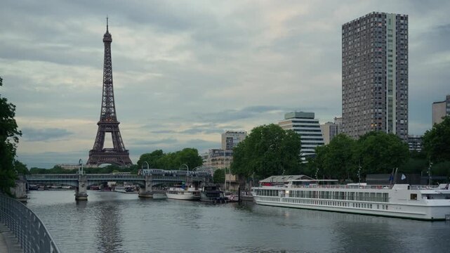 A passenger metro train crosses a river bridge near the Eiffel Tower and a docked cruise ship in Paris France. Metropolitan public transport infrastructure, urban commuter travel journey.