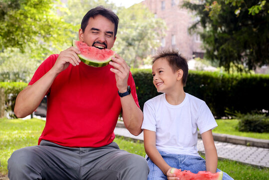 Playful father and son enjoying fresh watermelon in a sunny park. Happy family bonding, healthy summer snack, outdoor lifestyle, childhood joy, and wellness concept