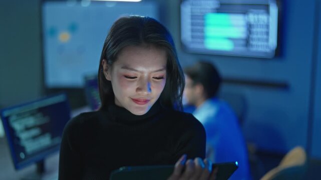 Young Asian woman smiling while working on tablet computer, male colleague and screens with code in background, tech team at night office.