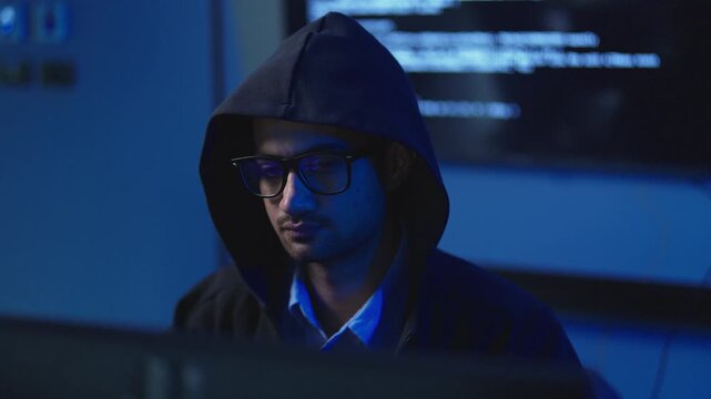 Young man wearing black hooded sweatshirt and eyeglasses in dark room with program code on screen behind, cyber crime and hacking concept.
