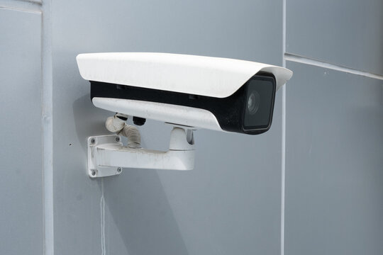 white surveillance camera is mounted on gray wall for monitoring. black surveillance camera watches public space. modern surveillance camera creates big brother control and privacy loss today.