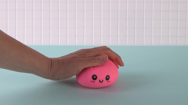 Hand presses and releases a pink squishy toy with a smiling face on a light blue surface, showcasing the toy's soft texture and playful design in a bright setting