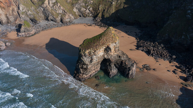 Aerial view of the Bedruthan Steps sea stack with greenery on top on a sandy beach with crashing waves and rugged cliffs in Padstow, Cornwall, United Kingdom.