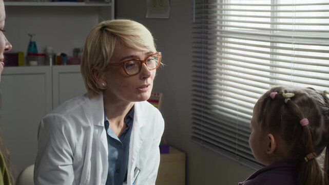 Waist up of Caucasian middle aged female pediatrician conducting neurological reflex test on Caucasian female child patient in clinic. Ideal for pediatric clinic promotions and healthcare marketing