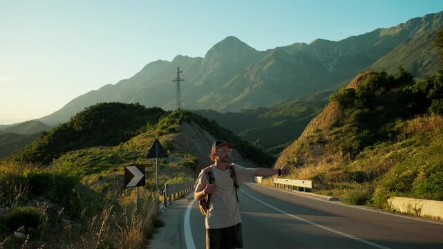 Handheld wide shot of male backpacker on mountain roadside signaling for ride, lifting cap to flag down a car. Warm sunset light, open road and waiting pose highlight hitchhiking and urgency.
