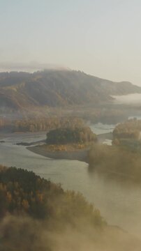 Aerial view of a misty river valley with forest and mountain ridges at sunrise. Soft morning fog drifts above trees and water, creating a peaceful natural landscape with atmospheric light and scenic