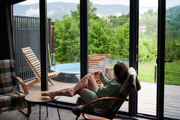 Obraz premium Woman relaxing in armchair with legs on table, enjoying scenic mountain view through large window, modern cozy cabin interior with terrace, outdoor hot tub and wooden lounge chairs