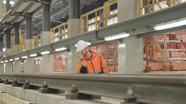 Construction worker communicates via radio with Electric Train operations within dark warehouse environment featuring steel beams and rail tracks equipment for industrial logistics management.