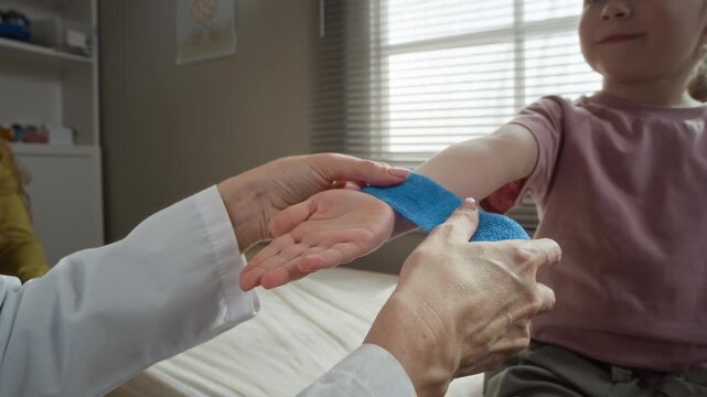 Closeup of female pediatrician wrapping blue elastic bandage around wrist of smiling Caucasian child girl in clinic. Clip is perfect for pediatric healthcare marketing and medical insurance promos